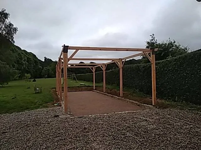 Pergola bois — Structure en bois brut sur sol stabilisé, au fond d'un jardin.