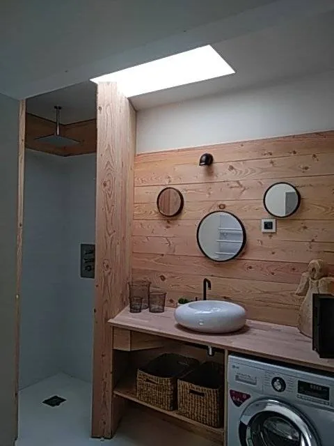 Salle de bains bois et blanc — Lambris naturel, vasque à poser, miroirs ronds, lave-linge intégré.